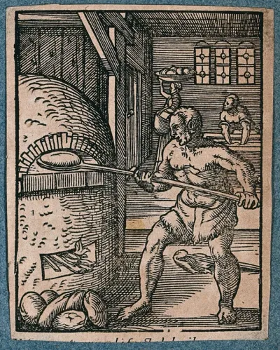 A baker is loading uncooked dough into an oven, as baked loaves are carried away by a woman