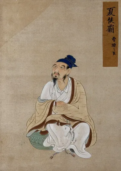 A Chinese figure, seated, wearing beige and white robes and blue hat black hat