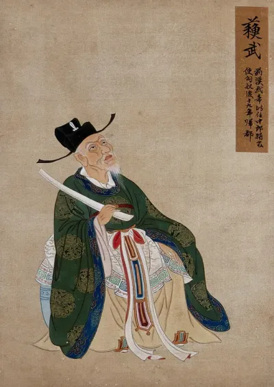 A Chinese figure, seated, wearing green robes richly decorated with lotus flower designs in gold thread with blue border and black hat