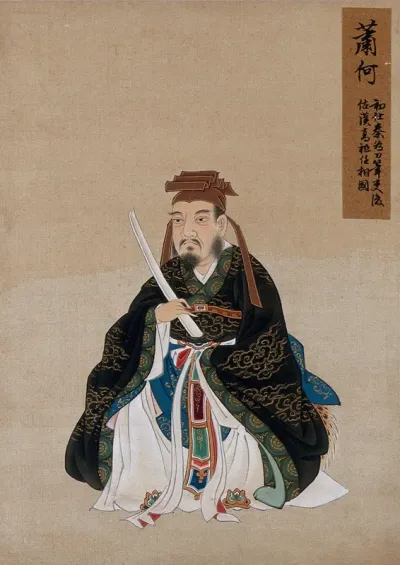 A Chinese figure wearing black silk robes with a green border, richly decorated with cloud scroll designs in gold thread