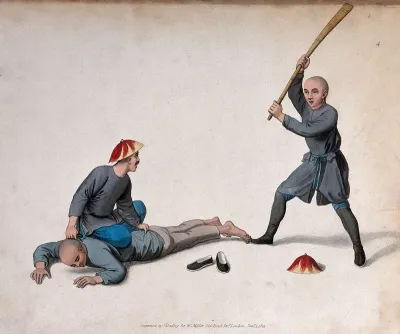 A Chinese man is held on the ground by a man while another is hitting him with a wooden stick