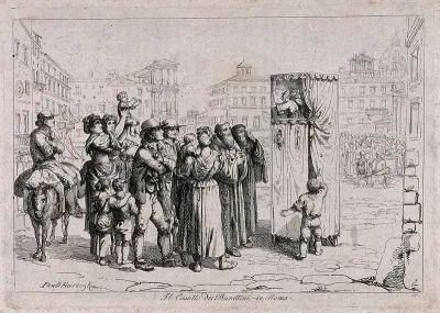 A crowd of people have gathered in the street to watch a Punch and Judy show, a mother has lifted her child up to see and another child pulls back the cloths to peep into the stand