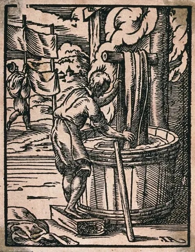 A dyer of cloth is dipping fabric into a large barrel as other sheets are hung up to dry