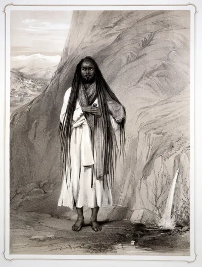 A Hindu man (sadhu) with long hair, cloak and walking stick stands by a rock from which water springs