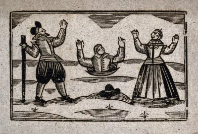 A man buried up to his waist holds his hands up above his head: a man and a woman stand on either side watching him in distress