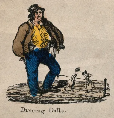A man has two dancing dolls on a board, controlled by a string attached to his leg