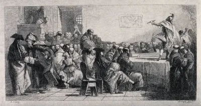 A man is performing on a table with a stick in his hand, people are sitting on chairs watching and an extra chair is being offered by a man in a hooded cape