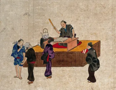 A man sits at a table on a platform reciting from a book, while three women and a man stand nearby
