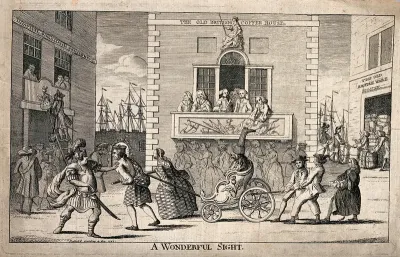 A procession with people pulling a dog in a carriage is going past a crowd of people on a balcony