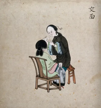 A standing woman preparing to remove facial hair from seated woman using a piece of string
