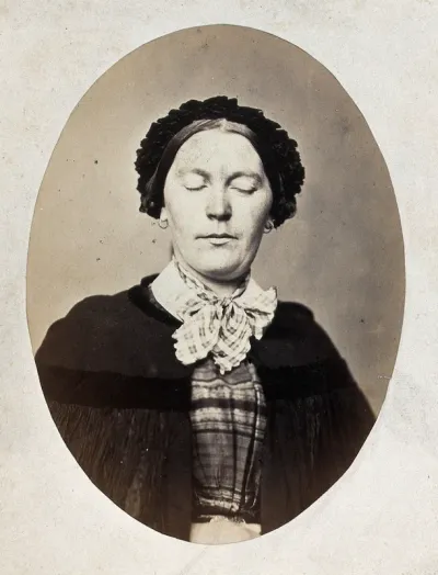 A woman's head and shoulders; her eyes are closed and her mouth shows a slight grimace