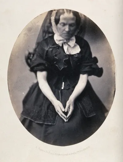 A woman, seated, wearing a black dress and veil; her hands are in her lap as though she is praying