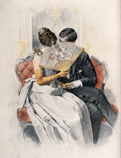 A young loving couple sit on a sofa holding hands while she coyly holds a fan up to her face