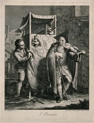 A young man plays a violin in front of a Punch and Judy stand with the puppeteer nearby