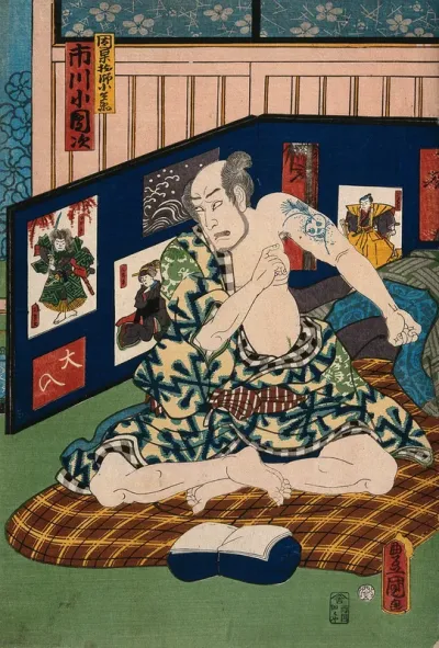 Actor Ichikawa Kodanji seated on bedding revealing a tattoo of a skull amid grasses on his left arm