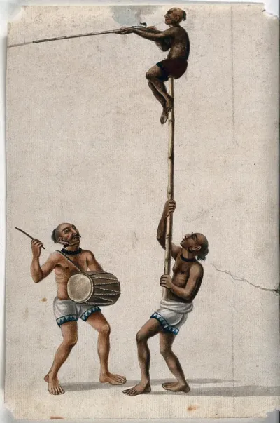 An acrobat climbing a pole held by another man while a musician drums out a beat