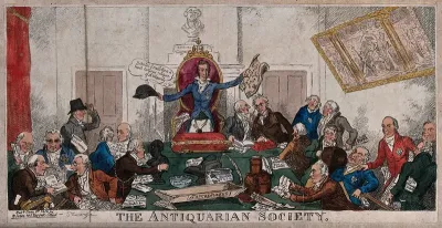 Antiquaries meeting: a man standing at the head of a table holds up a hat in one hand and a sheaf of papers in the other for the rest of the company to see; the table has busts and pots on it and the men are inspecting various items